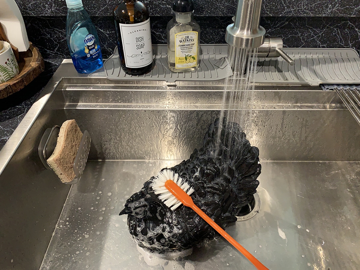 Black resin chicken getting a bath in the kitchen sink with a scrub brush.